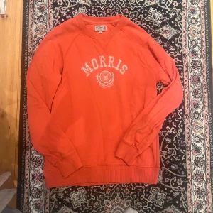Orange sweatshirt från Morris, M - Säljer en orange sweatshirt från Morris i storlek M. Tröjan har rund halsringning, ribbade muddar och tryckt logga med universitetsstil framtill. Perfekt för dig som gillar klassisk och sportig stil.
