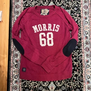 Mörkröd stickad tröja Morris M - Snygg mörkröd stickad tröja från Morris med vita bokstäver och siffran 68 framtill. Tröjan har svarta armbågslappar, ribbade muddar och rund hals. Perfekt för dig som gillar sportig och klassisk stil.