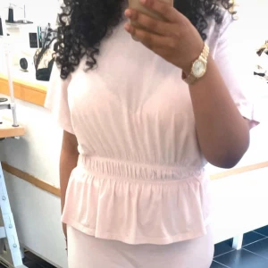 Ljusrosa kort klänning från 157 - Supersöt ljusrosa topp från 157 rynkad i midjan och volang nedtill. Toppen har korta ärmar och är gjord i mjuk bomull, perfekt för en chill och feminin look. Enkel och stilren design som passar till många tillfällen.