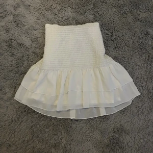 Vit volangkjol - Supersöt vit kortkjol med smockad midja och dubbla volanger nertill. Kjolen har en luftig och lätt känsla, perfekt för varma dagar. Den är figurnära upptill och faller snyggt med volangerna. Passar perfekt för en somrig look. Passar storlek xs-m.