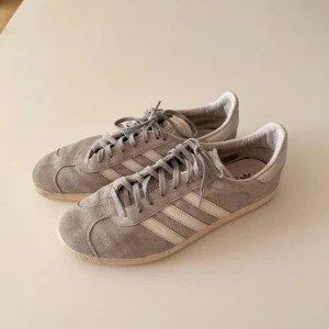 Adidas Gazelle grå mockasneakers - Grå Adidas Gazelle sneakers i mocka med klassiska vita ränder på sidorna och vit sula. Skorna har snörning och rund tå, samt diskret guldfärgad Gazelle-text på sidan. Perfekta för en clean och stilren look.