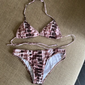Rosa och brun bikini med knyt - Säljer en snygg bikini i rosa och bruna toner med unikt mönster. Överdelen är trekantsmodell med knyt både i nacken och ryggen. Underdelen har klassisk bikini-passform. Perfekt för stranden eller poolen!