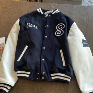 Blå och vit bomberjacka - Stake (Nypris 4000kr) - En oanvänd exklusiv bomberjacka från Stake i mörkblått med vita ärmar och detaljer. Jackan har broderad logga och stort S på bröstet, quiltat foder och ribbade muddar med vita ränder. Yttermaterialet är ull och ärmarna i skinn, med knappar framtill och två fickor. Limited Edition - Drake & Stake 