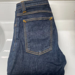 Mörkblå jeans från Nudie Jeans - Snygga mörkblå jeans från Nudie Jeans med klassisk desig och sömmar i gult. Jeansen har slim fit passform och är tillverkade i slitstarkt denim av bomull. Perfekta för dig som gillar en grisch och stilren look.