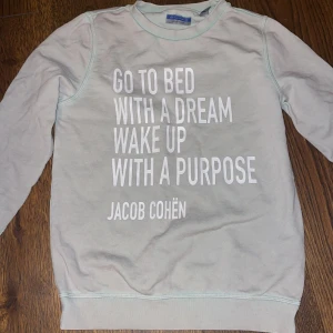 Ljusgrå sweatshirt från Jacob Cohën - Ljusgrå sweatshirt från Jacob Cohën med vit text på bröstet. Tröjan har rund halsringning, långa ärmar och ribbade muddar. Tillverkad i mjuk bomull, perfekt för chill dagar eller när du vill ha en clean look.