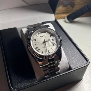 Seiko Day-Date Mod - Helt nybyggd Seiko Day-Date Mod  Specifikationer: Storlek: 39mm Urverk: Miyota 8285 (Automatiskt)  Material: 904L Rostfritt Stål Glas: Sapphire Glas Datumvisare och Dag visare Boett: Räfflad Bezel Tavla: Vit med Romerska Siffror Armband: Presidential och Justerbart  Krona: Screw Down Vattentålighet: 30 Meter Klocklåda ingår