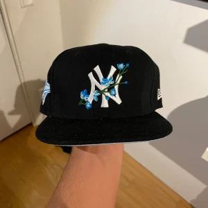 New Era Yankees keps med blommor - Har tre till färger som jag säljer då jag råka köpa fel stolek. Helt nya.Snygg svart New Era 59FIFTY Yankees keps med broderat NY-logga och blå blommor framtill. På sidan finns ett blått World Series 1996-märke. Kepsen är i 100% polyester och har klassisk rak skärm.