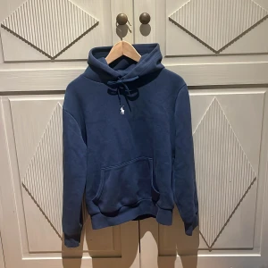 Blå hoodie från Polo Ralph Lauren - Säljer en snygg blå hoodie från Polo Ralph Lauren med klassisk vit logga på bröstet. Tröjan har huva med snörning, känguruficka och ribbade muddar. Perfekt för dig som gillar stilren och bekväm streetwear.