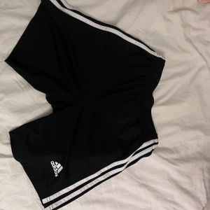 Svarta Adidas shorts med vita ränder - Svarta Adidas shorts med klassiska vita ränder längs sidorna och Adidas-logga framtill. Tillverkade i lätt och skönt syntetmaterial, perfekt för träning eller sport. Elastisk midja för bekväm passform. Modell Primegreen.