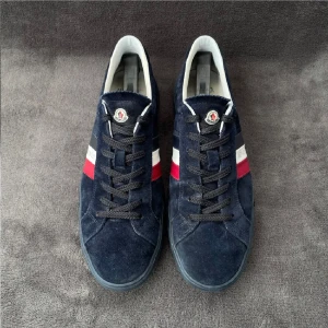 Moncler sneakers i mörkblå mocka - Snygga sneakers från Moncler i mörkblå mocka med klassiska röd och vit rand på sidan och Moncler-logga på plösen. Skorna har svarta snören, vit insida och platt sula. Perfekta för dig som vill ha en stilren och exklusiv look.