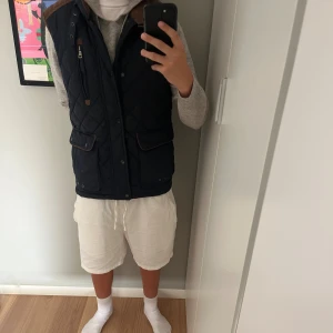 Mörkblå quiltad väst från Fenix Trading - Quiltad mörkblå väst med bruna detaljer från Fenix Trading. Västen har hög krage, dragkedja och tryckknappar framtill, samt två stora fickor med lock och en bröstficka med dragkedja. Snyggt rutmönster och kontrast på axlarna ger en cool look.