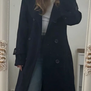 Mörkblå kappa med stora slag - Snygg mörkblå kappa från H&M med dubbelknäppning och stora slag. Jackan har långa ärmar med dekorativa knappar vid ärmsluten och en klassisk, rak passform. Perfekt för dig som vill ha en stilren och tidlös look under kyligare dagar.