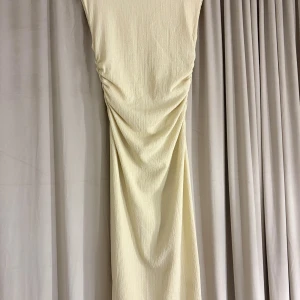 Ljusbeige långklänning med rynk - En elegant långklänning i ljusbeige med snygga rynkade detaljer längs sidorna. Klänningen har en mjukt texturerad yta och faller snyggt längs kroppen. Perfekt för dig som gillar stilrena och trendiga plagg med en twist.