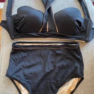 Dagi Svart bikini med guldiga detaljer  - Snygg svart bikini från Dagi med vadderad bygel-bh och hög midja på underdelen. Guldiga detaljer och insida med glittrigt leopardmönster ger en lyxig känsla. Materialet är stretchigt och bekvämt, perfekt för stranden eller poolen. Använt endast 1 gång. 