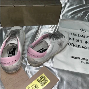 Golden Goose sneakers med rosa detaljer - Golden Goose sneakers i vitt skinn med grå snörning, silverfärgad stjärndetalj på sidan och ljusrosa hälparti. Snyggt slitna detaljer och platt sula. Perfekta för dig som gillar en cool och avslappnad stil.