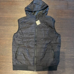 Svart quiltad dunväst från J.STYLE - Svart dunväst från J.STYLE med huva och quiltad design. Västen har två sidofickor med dragkedja och hel dragkedja framtill. Materialet är syntet och polyester, vilket gör den lätt och smidig. Perfekt för lager-på-lager under höst och vinter. Priset går att diskutera.