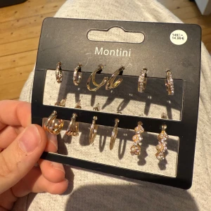 Montini örhängen multipack guld - Säljer tre olika Montini multipack med örhängen i guldton. Seten innehåller studs och hängande örhängen med glittriga stenar och pärlor. Vissa har randig och vågig design, andra är dekorerade med gnistrande detaljer och pärlor. Perfekt för att mixa och matcha till din stil. 10kr per par