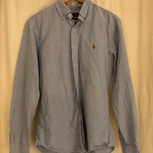 Blå skjorta Polo Ralph Lauren slim fit - Snygg ljusblå skjorta från Polo Ralph Lauren i slim fit med klassisk button-down krage och broderad logga på bröstet. Skjortan är långärmad och har knappar framtill. Perfekt för dig som gillar stilrena och tidlösa plagg.