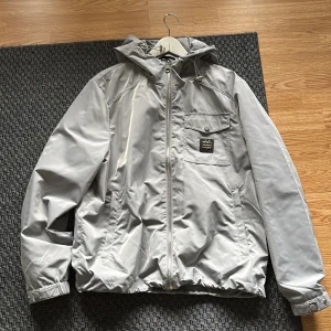 Ljusgrå vindjacka från Bvaldi, M - Snygg ljusgrå vindjacka från Bvaldi med huva och dragkedja framtill. Jackan har en bröstficka med lock och metallogo, samt två sidofickor. Justerbara muddar med tryckknappar och elastisk nederkant. Perfekt för blåsiga och regniga dagar. Köpt från bvaldis hemsida för ett tag sen men har inte kommit så mycket till användning så den är i princip ny.