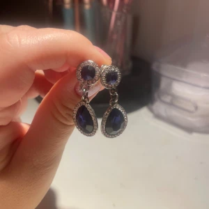 Glittriga blå droppformade örhängen - Snygga örhängen med droppformade mörkblå stenar och glittriga stenar runt om. Örhängena har en silverfärgad bas och hängande design som ger extra bling. Perfekta för att lyfta din look med lite extra glam.