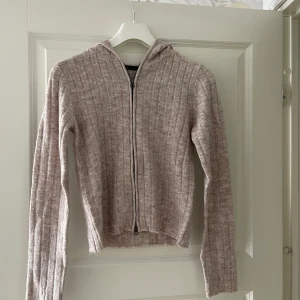 Beige ribbad hoodie med dragkedja - Mysig ribbstickad hoodie i beige med dragkedja framtill och huva. Tröjan har lång ärm och är tillverkad i ett mjukt stickat material som passar perfekt för kyligare dagar. Enkel och stilren design som är lätt att matcha.