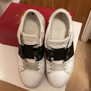 Valentino Garavani Open sneakers vit/svart - Snygga Valentino Garavani Open sneakers i vitt skinn med bred svart rem över vristen. Klassisk rund tå, platt sula och coola nitar baktill på sulan. Skorna har snörning och guldiga logodetaljer på plösen. Perfekta för dig som gillar exklusiva grisch-vibbar. Ingår även skolåda, skopåse och ett valentino garavani kort. Notera liten defekt på vänster sida av skon