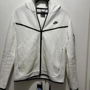 Vit hoodie från Nike med svarta detaljer - Nike hoodie i vit med svarta kontrastdetaljer och dragkedja. Tröjan har huva, långärmad design och en cool svart ficka på ärmen. Tröjan är helt ny har inte aldrig använt den. Tröjan är köpt från Nike men kom med fel storlek så det gick inte att göra retur. Tröjan är köpt för 1400kr och har kvar allt som kommer med den. Pris kan diskuteras!