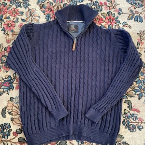 Mörkblå stickad tröja från Indicode - Säljer en mörkblå stickad tröja från Indicode i storlek XL. Tröjan har ett snyggt kabelstickat mönster, hög krage och half zip-dragkedja framtill. Perfekt för dig som gillar klassisk stil med en modern twist.