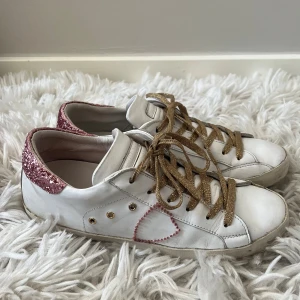 Philippe Model skor - Snygga vita Philippe Model sneakers i skinn med guldiga snören, rosa glitter på hälen och ett broderat hjärta på sidan. Perfekta för dig som vill sticka ut med lite extra bling. Köpta på sellpy. 