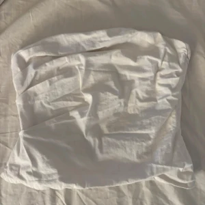 Vit bandeau tubtopp från Zara - Säljer en stilren vit bandeau tubtopp från Zara i bomull. Toppen är axelbandslös och har dragkedja baktill för enkel på- och avtagning. Perfekt att matcha med jeans eller kjol för en clean look.