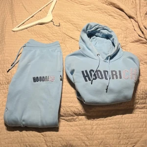 Ljusblå HOODRICH hoodie med brodyr - Säljer en ljusblå hoodie från HOODRICH med broderad logga i svart och silver på bröstet. Hoodien har huva med snörning, magficka och långa ärmar. Mjuk och skön, perfekt för chill dagar.