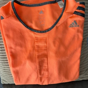 Orange Adidas träningstshirt dam L - •Sportig orange t-shirt från Adidas med grå detaljer och klassiska ränder på axlarna.  •Använd 1 gång. Som ny. Tvättad. •Tillverkad i lätt och ventilerande Climalite-polyester, perfekt för träning.  •Kortärmad och figurnära passform med rund halsringning.  🚭Djurfritt och rökfritt hem