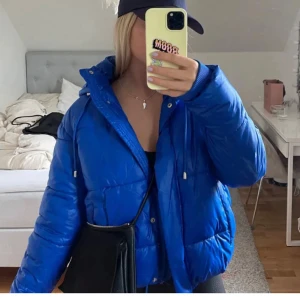 Blå pufferjacka med huva - Säljer en riktigt snygg blå pufferjacka med huva och dragsko. Jackan har en cool, oversized passform och är perfekt för kalla dagar. Materialet är glansigt och känns skönt mot kroppen. Jackan har dragkedja framtill och ribbade muddar vid ärmslut.