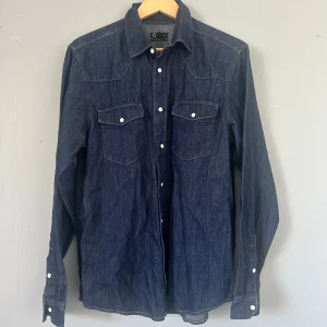 Mörkblå jeansskjorta  - Snygg mörkblå jeansskjorta  med klassisk western-feeling. Skjortan har vita tryckknappar, två bröstfickor med lock och lång ärm. Perfekt för dig som gillar en stilren och tidlös look med denim.