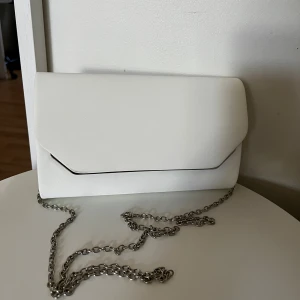 Vit kuvertväska med kedja - Stilren vit kuvertväska med silverfärgad kedja som axelrem. Väskan har en minimalistisk design med raka linjer och ett elegant kuvertlock. Eftersom att man kan lägga in kedjan i väskan så är den Perfekt för till exempel bal ☀️