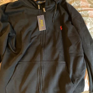 Svart hoodie från Polo Ralph Lauren  - Svart hoodie från Polo Ralph Lauren i storlek L. Klassisk design med dragkedja, huva med snörning och känguruficka. Broderad röd logga på bröstet. Perfekt för en chill och stilren look.