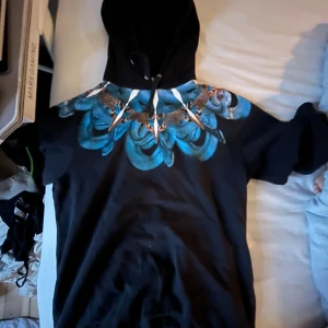 Marcelo Burlon hoodie med ormmönster - Svart hoodie från Marcelo Burlon med unikt blått och brunt ormmönster över axlarna. Tröjan har huva med snörning och är tillverkad i mjukt material.