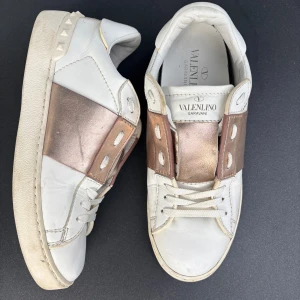 Valentino Garavani sneakers med guld - Snygga vita sneakers från Valentino Garavani med bred metallic guldrem över vristen och coola nitar på hälen. Skorna är i skinn och har klassisk snörning framtill. Perfekta för dig som vill sticka ut med en lyxig touch.