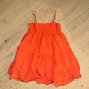 Korallfärgad klänning från Ghanda - Superfin orange kortklänning från Ghanda (Australien) med smock upptill och tunna axelband. Klänningen har volangig kjol och är gjord i ett lätt, skrynkligt bomullstyg som känns perfekt för varma dagar. Köpt för 350kr, säljer för 199