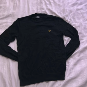 Svart stickad tröja från Lyle & Scott - Svart stickad tröja från Lyle & Scott med klassisk rund hals och gul broderad örn på bröstet. Tröjan har lång ärm och ribbade muddar vid ärmslut och nederkant. Perfekt för lager-på-lager och snygg till jeans.