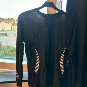 Svart långärmad träningströja Helly Hansen - Svart långärmad träningströja från Helly Hansen med vita randiga detaljer på sidorna och ärmarna. Tillverkad i lätt och ventilerande polypropylen, perfekt för aktiva dagar. Snygg passform och sportig look.