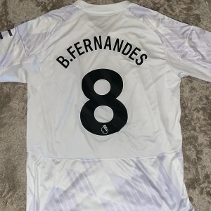 Manchester United Fernandes tröja M - Manchester United bortatröja från Adidas med B. Fernandes och nummer 8 på ryggen. Tröjan är vit med ljuslila grafiskt mönster, svarta detaljer och sponsorlogga på bröstet. Tillverkad i lätt och ventilerande polyester med klassisk V-ringning och korta ärmar.