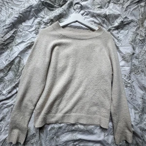 Beige stickad tröja från Vero Moda - Mysig beige stickad tröja från Vero Moda med bred ribbad kant vid halsen och ärmsluten. Tröjan har en enkel och stilren design med lång ärm och relaxed fit, perfekt för lager-på-lager under kyliga dagar.