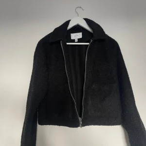 Svart teddyjacka från Vila, strl 36 - Mysig svart teddyjacka från Vila i storlek 36. Jackan har en klassisk krage, grov dragkedja framtill och är tillverkad i ett fluffigt material som ger en cool look. Perfekt för dig som gillar en enkel men trendig stil.
