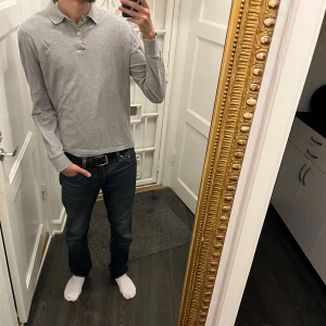 Grå långärmad pikétröja J.Lindeberg - Säljer en stilren grå långärmad pikétröja från J.Lindeberg. Tröjan har klassisk krage, knäppning med två knappar och diskret JL-logo på bröstet. Tillverkad i mjuk bomull som känns skön mot huden. Perfekt för en clean och avslappnad look.