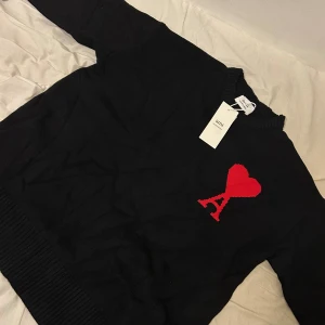 Svart stickad tröja från Ami Paris - Svart stickad tröja från Ami Paris med rund halsringning och ribbade muddar. På bröstet finns ett rött hjärta med bokstaven A, helt ny står L men är M