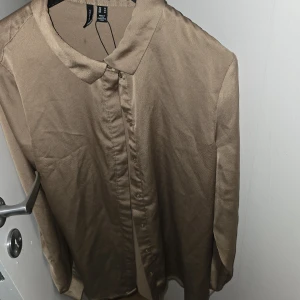 Gulig / Beige satinliknande skjorta / blus Vero Moda strl M - Guldig / beige skjorta från Vero Moda i satinliknande polyester. Klassisk krage, knäppning framtill och långa ärmar. Har en liten fläck baktill, (hår.säkert boatt få bort) i övrigt nyskick