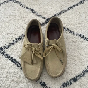 Beige mockaskor från Clarks Originals - Snygga beige mockaskor från Clarks Originals med klassisk design och rund tå. Skorna har snörning och är tillverkade i mjuk mocka med en platt sula. Perfekta för dig som gillar stilrena och tidlösa skor.