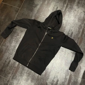 Svart hoodie från Lyle & Scott - Svart hoodie från Lyle & Scott med dragkedja och huva. Klassisk gul logga broderad på bröstet. Tröjan har långa ärmar, ribbade muddar och två fickor framtill. Perfekt för en chill och stilren look.