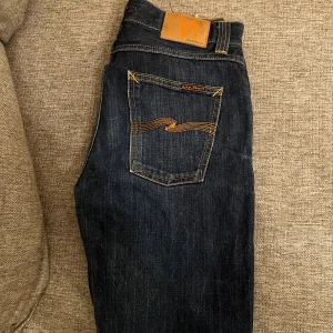 Mörkblå Nudie Jeans, storlek 30/32 - Säljer ett par mörkblå jeans från Nudie Jeans med klassisk orange söm på bakfickorna och läderpatch i midjan. Jeansen har rak passform och är tillverkade i slitstarkt denimtyg av bomull. Perfekta för dig som gillar stilrena och tidlösa jeans.